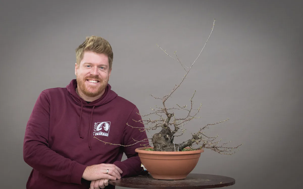 Lächelnder Teilnehmer neben einem unbelaubten Bonsai mit kräftigem Stammfuß und langem Spitzentrieb.