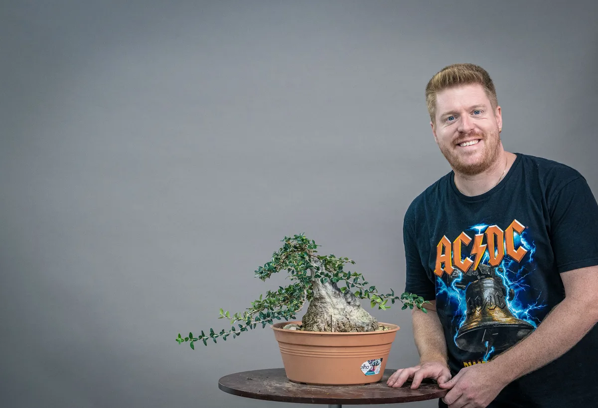 A workshop participant works on a small deciduous bonsai with a thick trunk and long lateral branches.